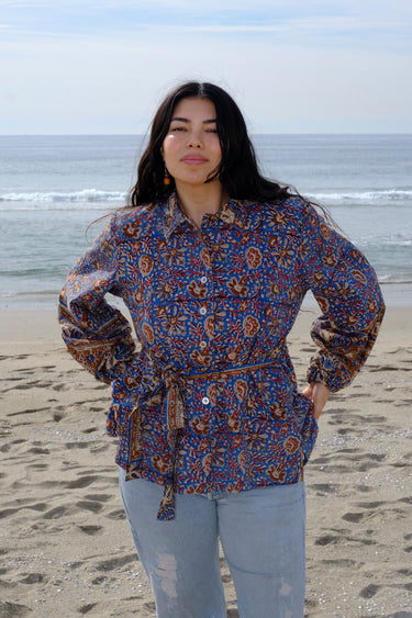 A woman wearing a blue floral tapestry jacket with big patch pockets and an elastic shirred sleeve cuff, standing by the beach. She is also wearing jeans.