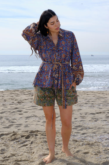 A woman wearing a blue floral tapestry jacket with big patch pockets and an elastic shirred sleeve cuff, standing by the beach. She is wearing coordinating shorts.
