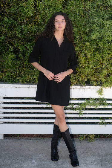 A woman standing in front of greenery wearing a black mini shirtdress with a collar, sleeves, and patch breast pockets, made of crinkle textured osnaburg cotton.