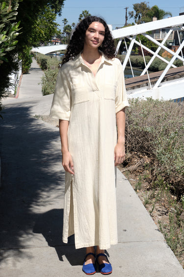 A woman standing on a pathway wearing an alabaster beige maxi shirtdress with a collar, sleeves, and a side slit, with a body made of crinkle textured osnaburg cotton.