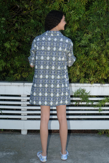 A person standing by a white fence with greenery wearing a mini shirt dress with a shirt collar and an "xo" Ikat pattern, back view.