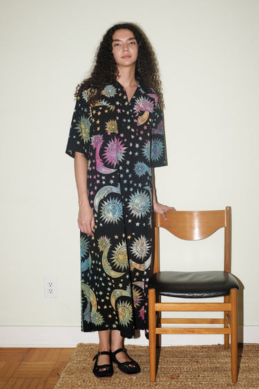 A woman inside standing next to a wooden chair, wearing a shirt dress caftan cut from a black ground tapestry printed with rainbow sun, moon and stars.