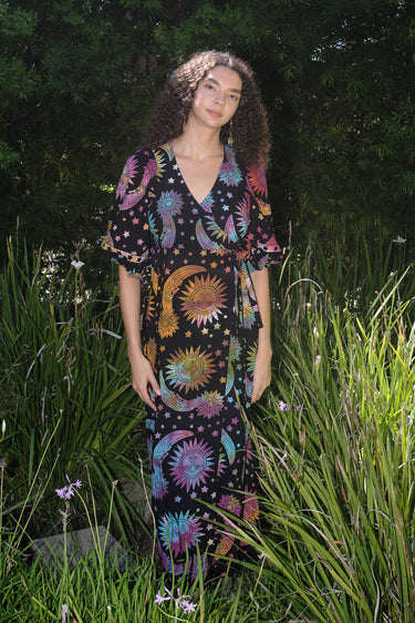 A woman standing in a garden wearing a multicolored maxi wrap dress with a tie-dye pattern and star shapes, featuring a wrap neckline and three-quarter sleeves.