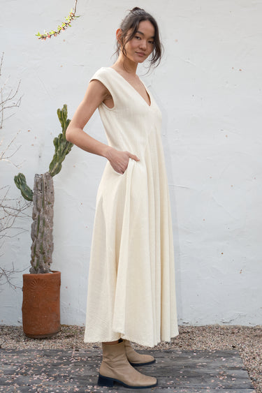 Woman in a long alabaster dress standing outdoors against a white wall with a cactus in the background.