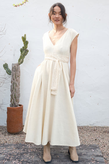 Woman in a long alabaster dress standing against a white wall with a cactus in the background.