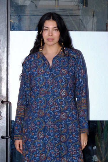 A woman wearing a midnight sky handloom tapestry button front shirt dress with a band collar and side panels, featuring a tie at the back waist, standing in front of a door.