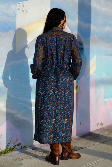 A woman wearing a midnight sky handloom tapestry button front shirt dress with a band collar and side panels, featuring a tie at the back waist, standing in front of a rainbow painted wall. Back View
