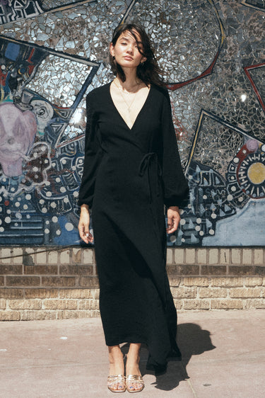 A woman standing in front of a colorful mosaic wall, wearing a black maxi dress with a wrap front, long full sleeves, and a bias quarter skirt.