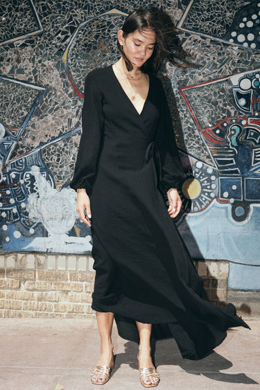 A woman standing in front of a colorful mosaic wall, wearing a black maxi dress with a wrap front, long full sleeves, and a bias quarter skirt.