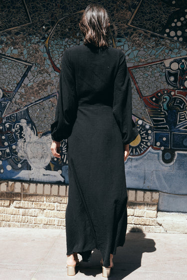 A woman standing in front of a colorful mosaic wall, wearing a black maxi dress with a wrap front, long full sleeves, and a bias quarter skirt, back view.