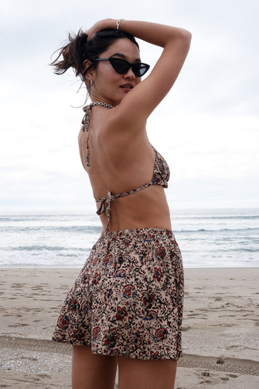 A person wearing sunglasses and a floral patterned woven swimsuit shorts, posing on a beach with the ocean in the background.