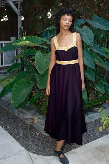 Woman in aNoire black dress with yellow accents standing in front of green foliage.