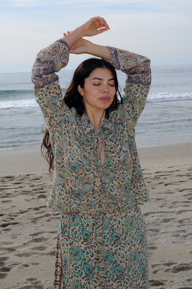 A woman standing at the beach wearing tan with green, turquoise flower tapestry shirt with bell sleeves and a matching skirt.