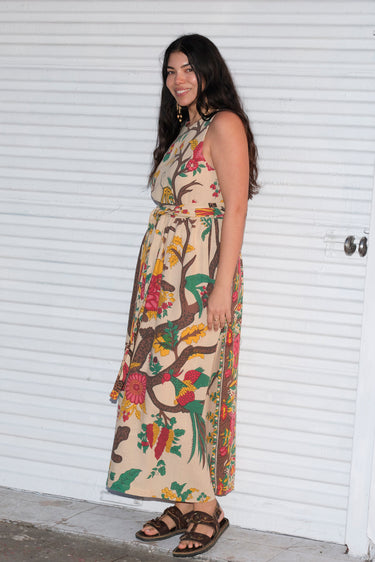 A woman wearing a beige maxi dress with a tree of life tapestry pattern, featuring wide ties and a slight empire waist. She is standing by a white roll up door.