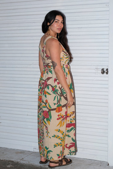 A woman wearing a beige maxi dress with a tree of life tapestry pattern, featuring wide ties and a slight empire waist. She is standing by a white roll up door. Side Back View