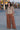 A woman standing at a beach boardwalk wearing a nutmeg brown trouser and a floral print blouse.