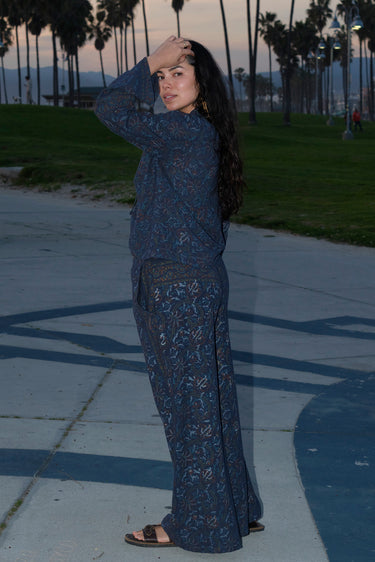 A woman standing at Venice Beach at dusk wearing dark blue midnight sky handloom tapestry low rise trouser and a matching shirt with bell sleeves. Side View.