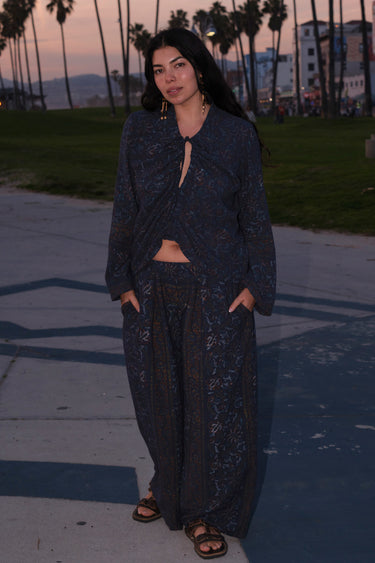 A woman standing at Venice Beach at dusk wearing dark blue midnight sky handloom tapestry low rise trouser and a matching shirt with bell sleeves.