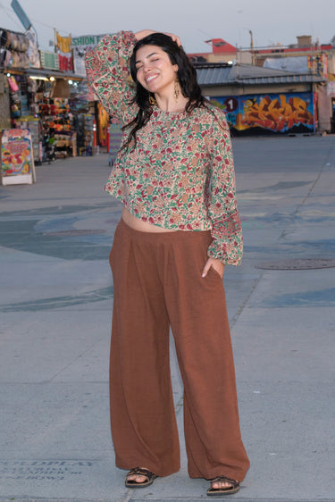 A woman standing at a beach boardwalk wearing a nutmeg brown trouser and a floral print blouse.