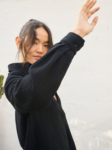 A woman wearing a black textured long-sleeve top with a buttoned cuff, posing with one arm raised against a light gray wall.