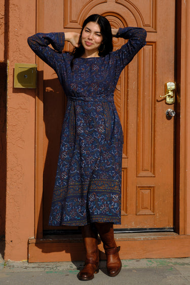A woman standing in front of a door, wearing a blue tapestry wrap dress with long bell sleeves and a quarter circle skirt, featuring patch pockets.