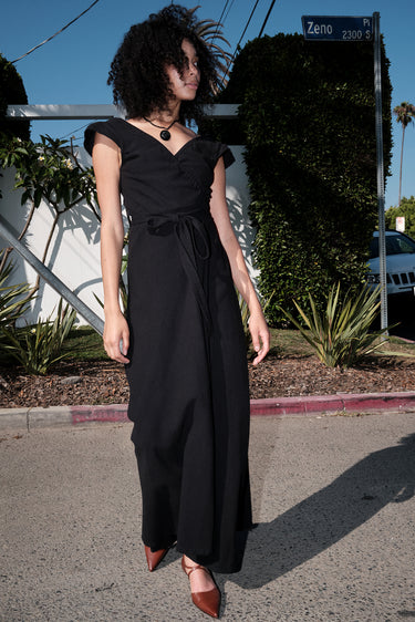 A woman standing outside wearing a long black maxi wrap dress with an empire waist and an elastic neckline.