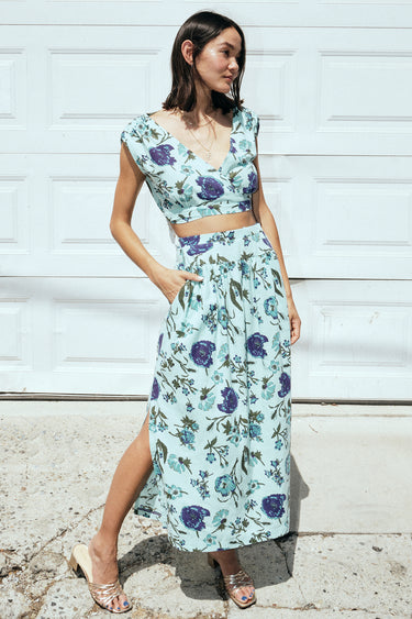 A woman standing outside by a white garage wearing an aqua ground floral tapestry skirt with a drop waist and side slits, paired with a matching cocoa top.