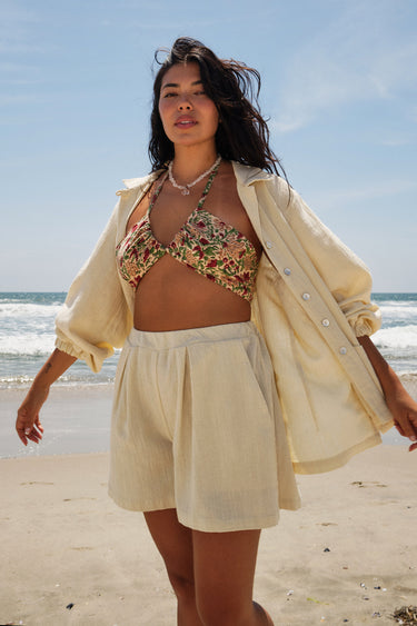 A woman standing on a beach wearing a floral tapestry bikini top and an alabaster trouser short, with a matching alabaster shirt jacket.