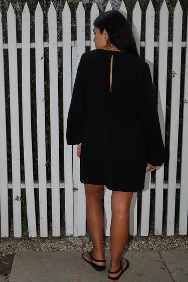 A woman standing in front of a white fence, wearing a black mini dress with a gathered neckline, long full sleeves, and a split back. The dress is made of a crinkle textured cotton fabric. Back View