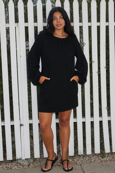 A woman standing in front of a white fence, wearing a black mini dress with a gathered neckline, long full sleeves, and a split back. The dress is made of a crinkle textured cotton fabric.
