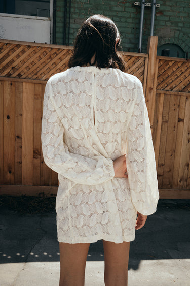 A woman standing outside by a brown wood fence wearing a lace mini dress, back view.