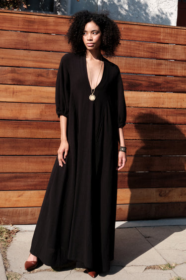 A woman standing outside by a brown wood fence wearing a maxi length black v neck caftan.