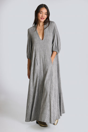 A woman standing in a white walled photo studio wearing a 3/4 sleeve maxi caftan cut from a salt and pepper textured cotton.