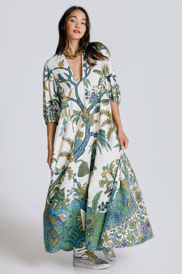 A model standing in a white photo studio wearing a maxi caftan, with three quarter balloon sleeves and pockets. The caftan is cut from a blue and green hand block printed tree of life tapestry