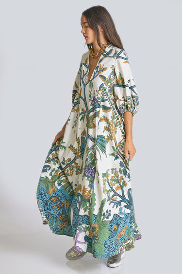 A model standing in a white photo studio wearing a maxi caftan, with three quarter balloon sleeves and pockets. The caftan is cut from a blue and green hand block printed tree of life tapestry