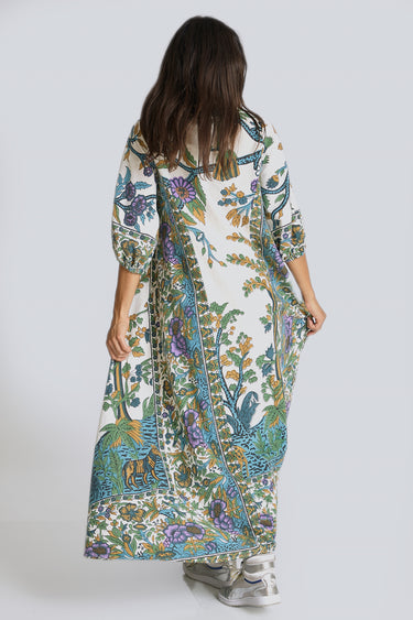 A model standing in a white photo studio wearing a maxi caftan, with three quarter balloon sleeves and pockets. The caftan is cut from a blue and green hand block printed tree of life tapestry, back view.