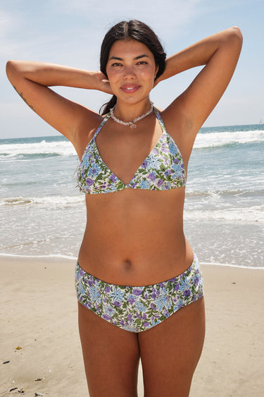 A woman at the beach wearing a white ground, lavender and blue floral tapestry triangle bikini top and matching bikini bottom.