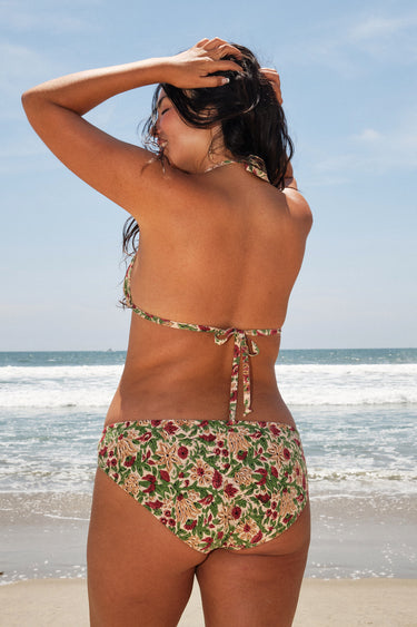 A woman at the beach wearing a beige ground, red and brown floral tapestry triangle bikini top and matching bikini bottom. Back View