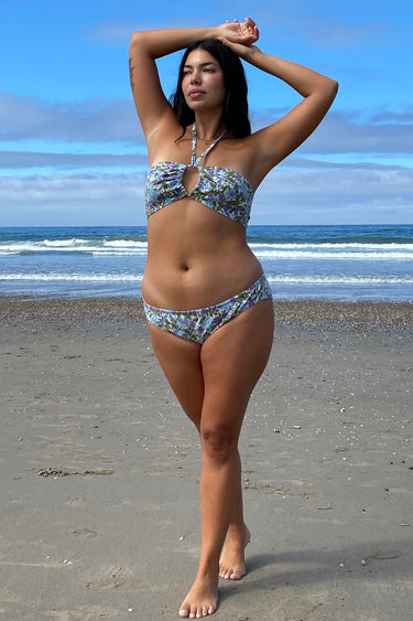 A woman at the beach wearing a white ground, lavender and blue floral tapestry bandeau bikini top and matching bikini bottom.