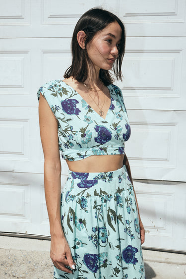 A woman wearing an aqua ground tapestry crop top and a matching skirt, standing in front of a white door.