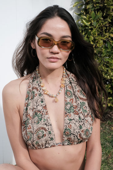 A woman standing outside by a white wall wearing a tan with black, brown and green paisley handloom tapestry halter bikini top.