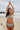 A woman on a beach wearing a white ground with lavender, blue and green floral tapestry bikini bottom with a high rise and matching bikini top.