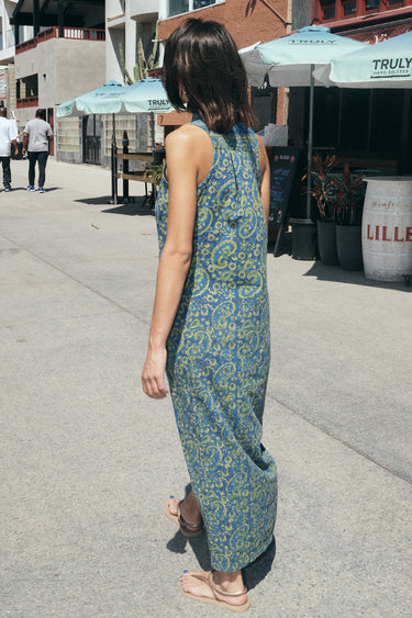 A woman on a beach boardwalk wearing a sheath dress made from a dark blue paisley tapestry, side back view.