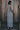 A model wearing a gray maxi shift dress with a front zipper and pockets, standing near a wooden fence. Back View