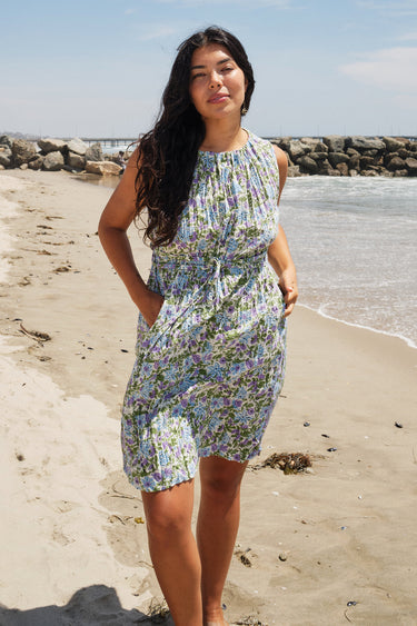 A woman standing on a beach wearing a knee-length floral dress with broomstick pleats, featuring an elastic neckline and a slit back. The dress is a white ground tapestry with blue and lavenders flowers.