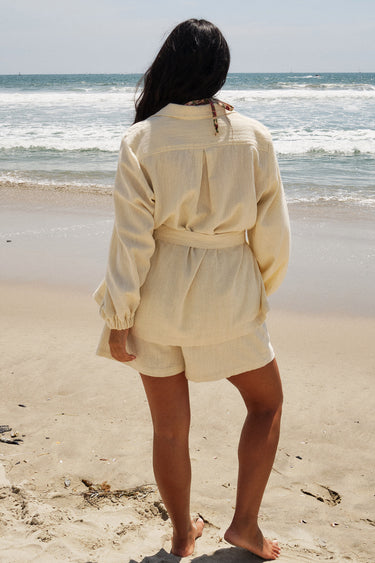 A woman standing on a beach wearing an alabaster trouser short with a matching alabaster shirt jacket. Back View