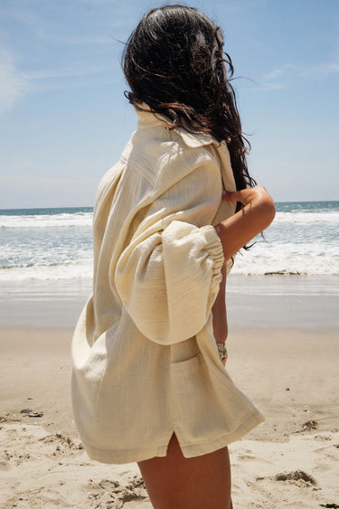 A woman standing on a beach wearing an alabaster unlined shirt jacket with big patch pockets and an elastic shirred sleeve cuff, side view.