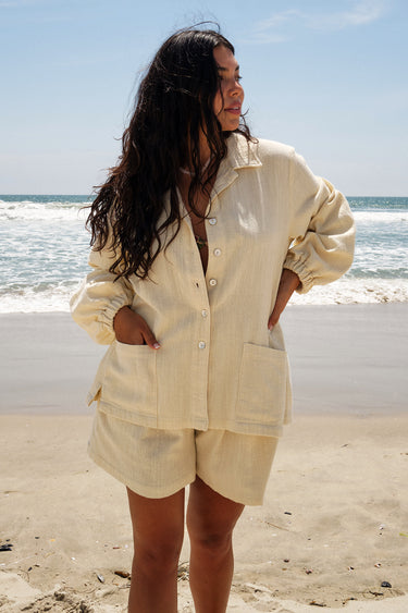 A woman standing on a beach wearing an alabaster unlined shirt jacket with big patch pockets and an elastic shirred sleeve cuff, with matching shorts.