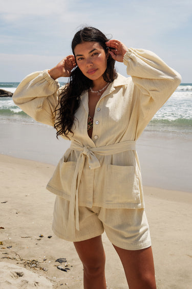 A woman standing on a beach wearing an alabaster unlined shirt jacket with big patch pockets and an elastic shirred sleeve cuff, with matching shorts.