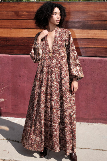 A woman standing by a red wall, outside wearing a long sleeve maxi caftan cut in a raisin floral tapestry.